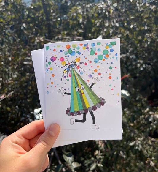 A hand holds a white greeting card outdoors in front of blurred green foliage. The card features a colorful illustration of a party hat character with a striped green and yellow cone, black limbs, and colorful confetti and dots on a white background.