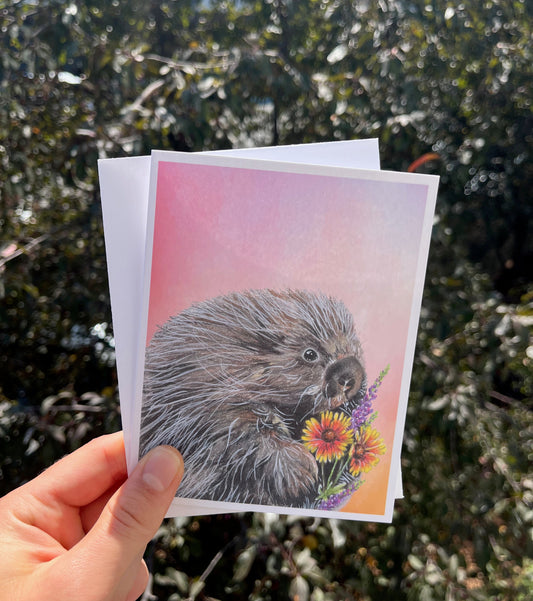A hand holds a printed art card featuring a detailed illustration of a hedgehog with flowers against a pink and orange background, with a blurred outdoor garden backdrop.