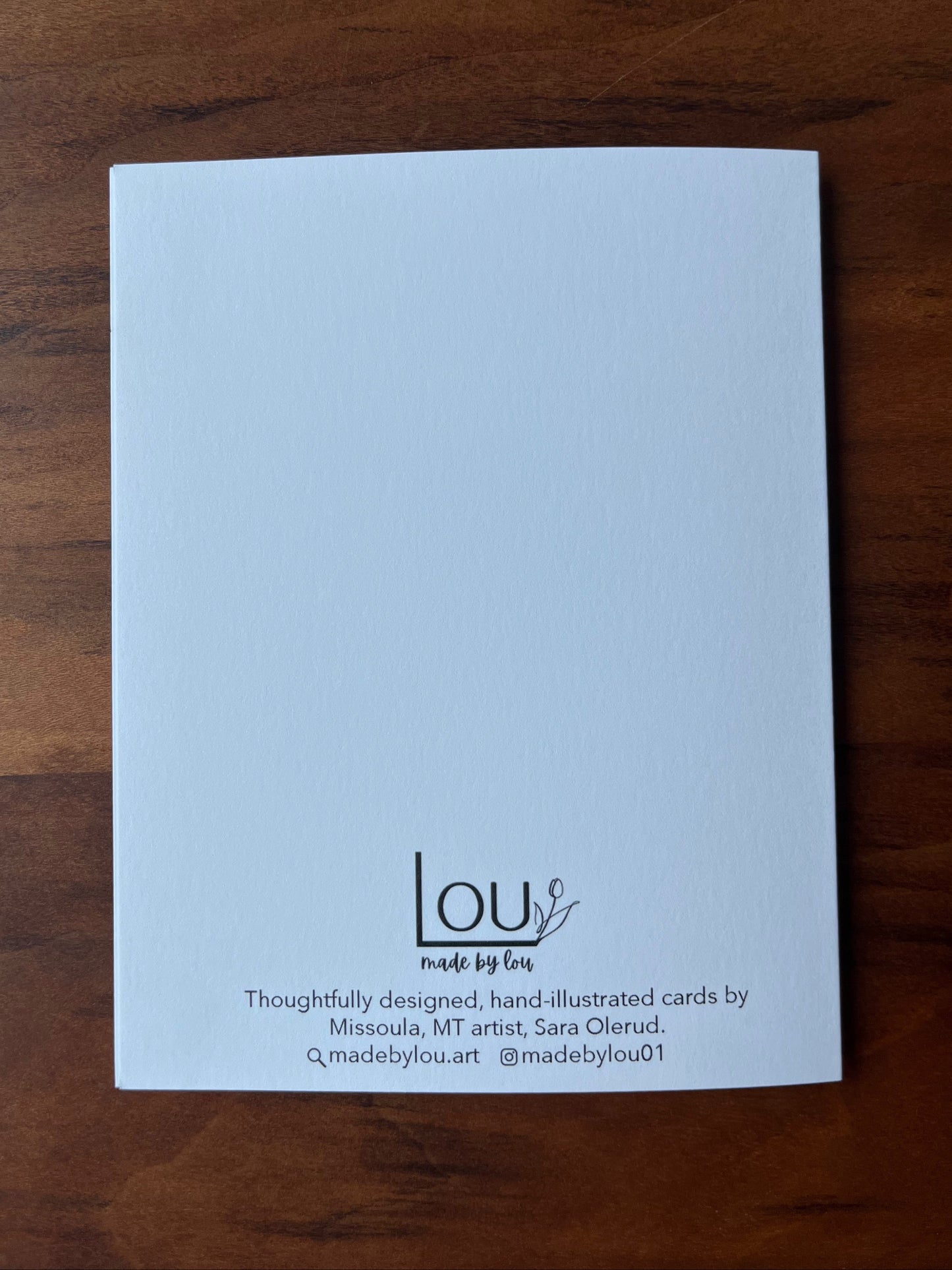 A white textured card with black printed text and logo at the bottom, placed on a dark brown wooden surface. The card is branded 'lou made by Lou'.