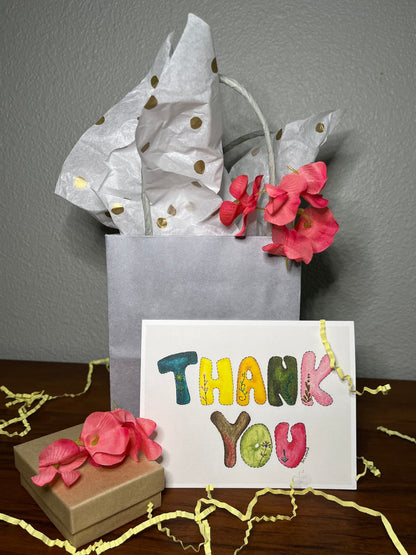 Gift bag with flowers, small box, and 'Thank You' card on a wooden surface.