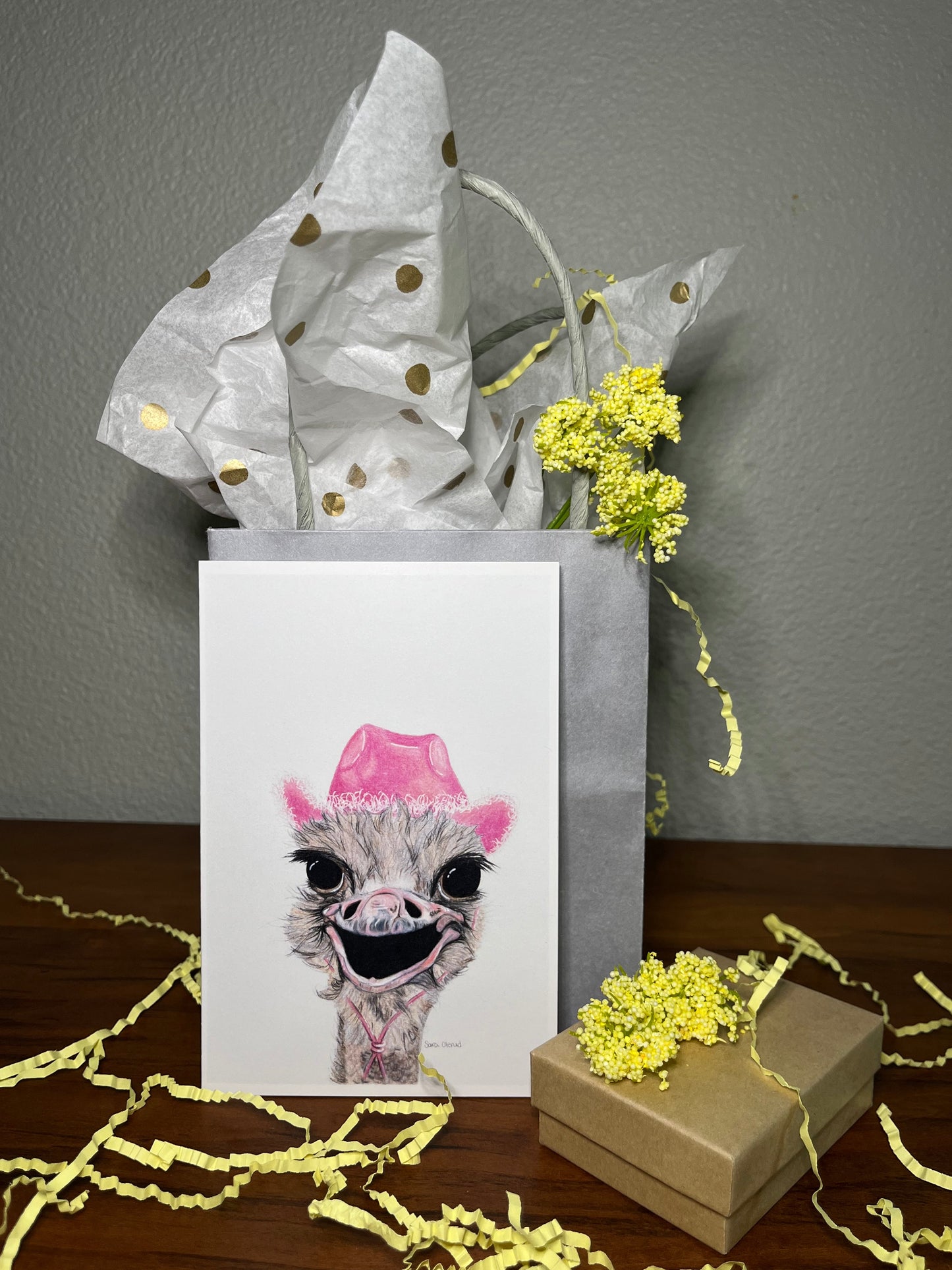 Gift box with a card featuring an ostrich wearing a pink hat, surrounded by yellow flowers on a wooden surface.