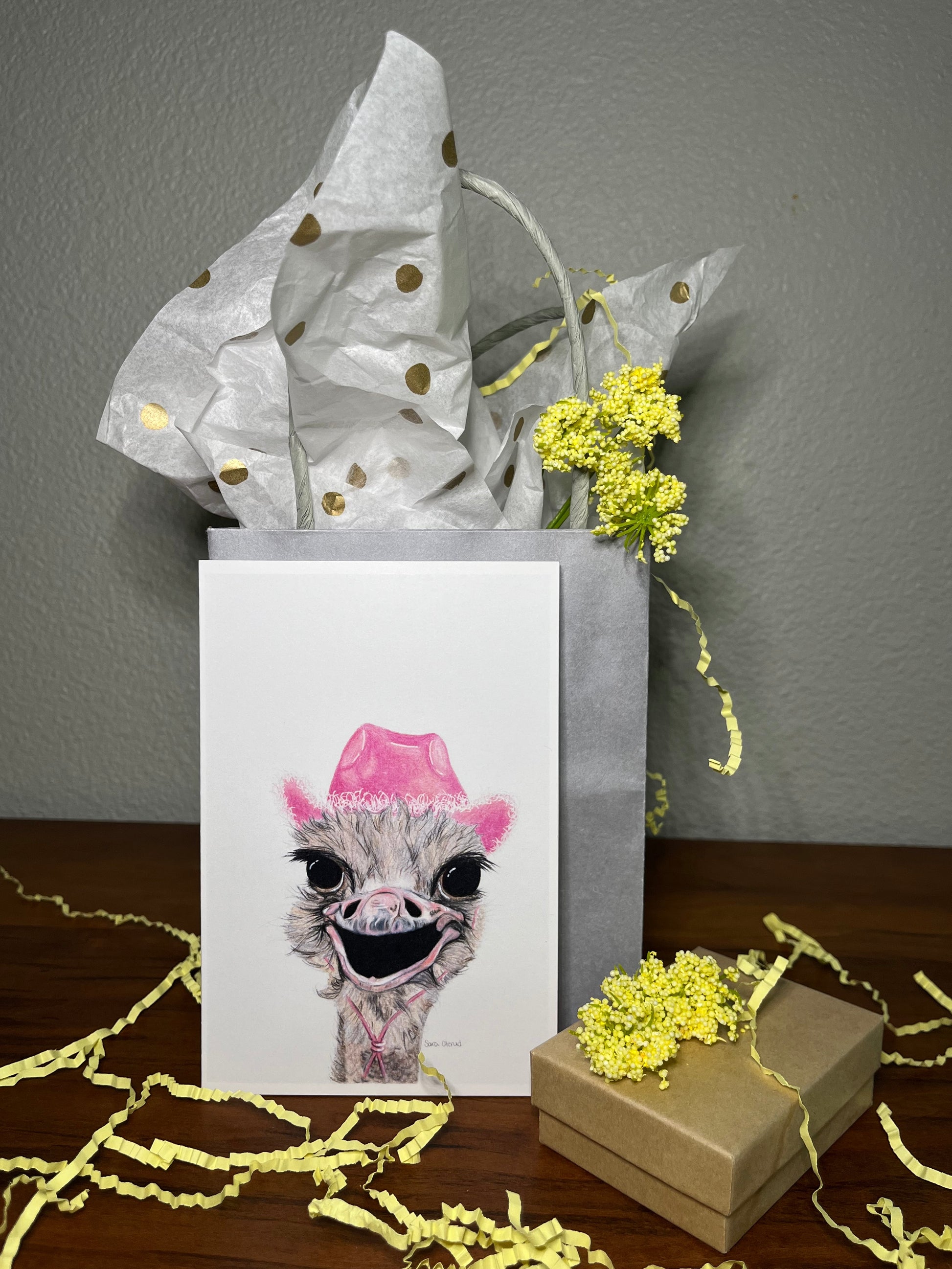 Gift box with a card featuring an ostrich wearing a pink hat, surrounded by yellow flowers on a wooden surface.