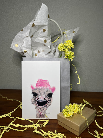 Gift box with a card featuring an ostrich wearing a pink hat, surrounded by yellow flowers on a wooden surface.