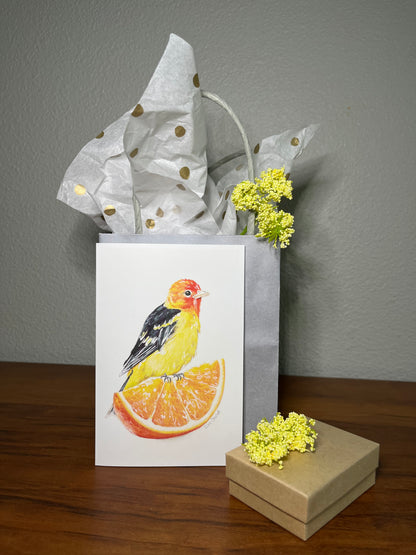 Gift bag with a bird design on an orange slice, surrounded by yellow flowers on a wooden surface.