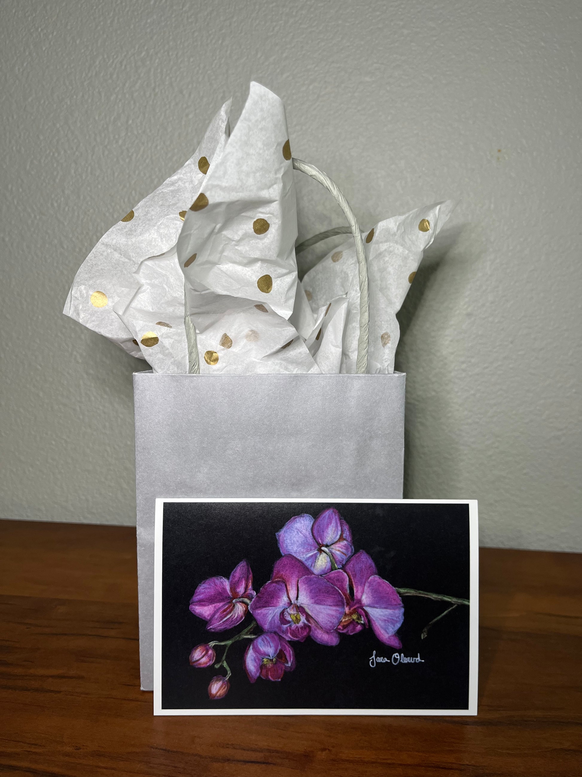Gift bag with white paper and gold dots, featuring a card with purple orchid illustration on a wooden surface.