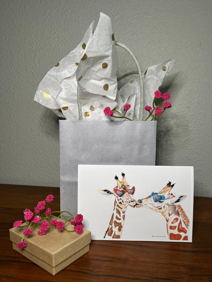Greeting card with giraffe illustration, wrapped gift, and small box on a wooden surface.