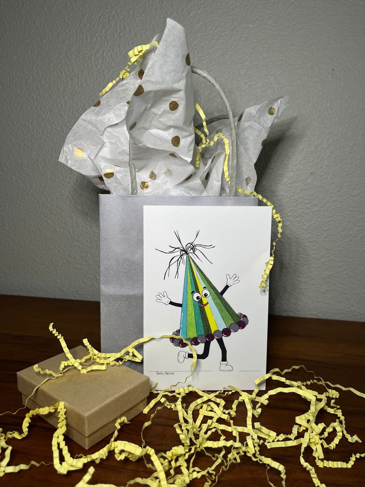 Gift bag with a card featuring a colorful character, surrounded by yellow paper shreds on a wooden surface.