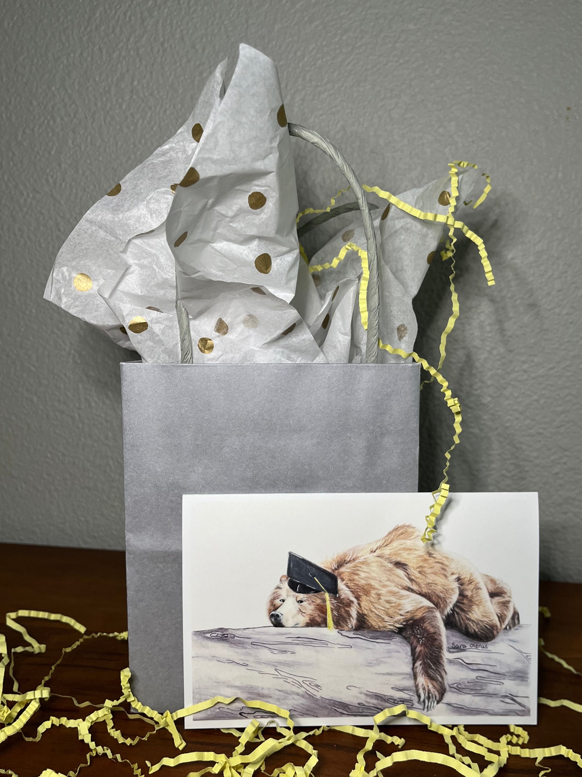Gift bag with gold polka dots and a card featuring a bear illustration on a wooden surface.