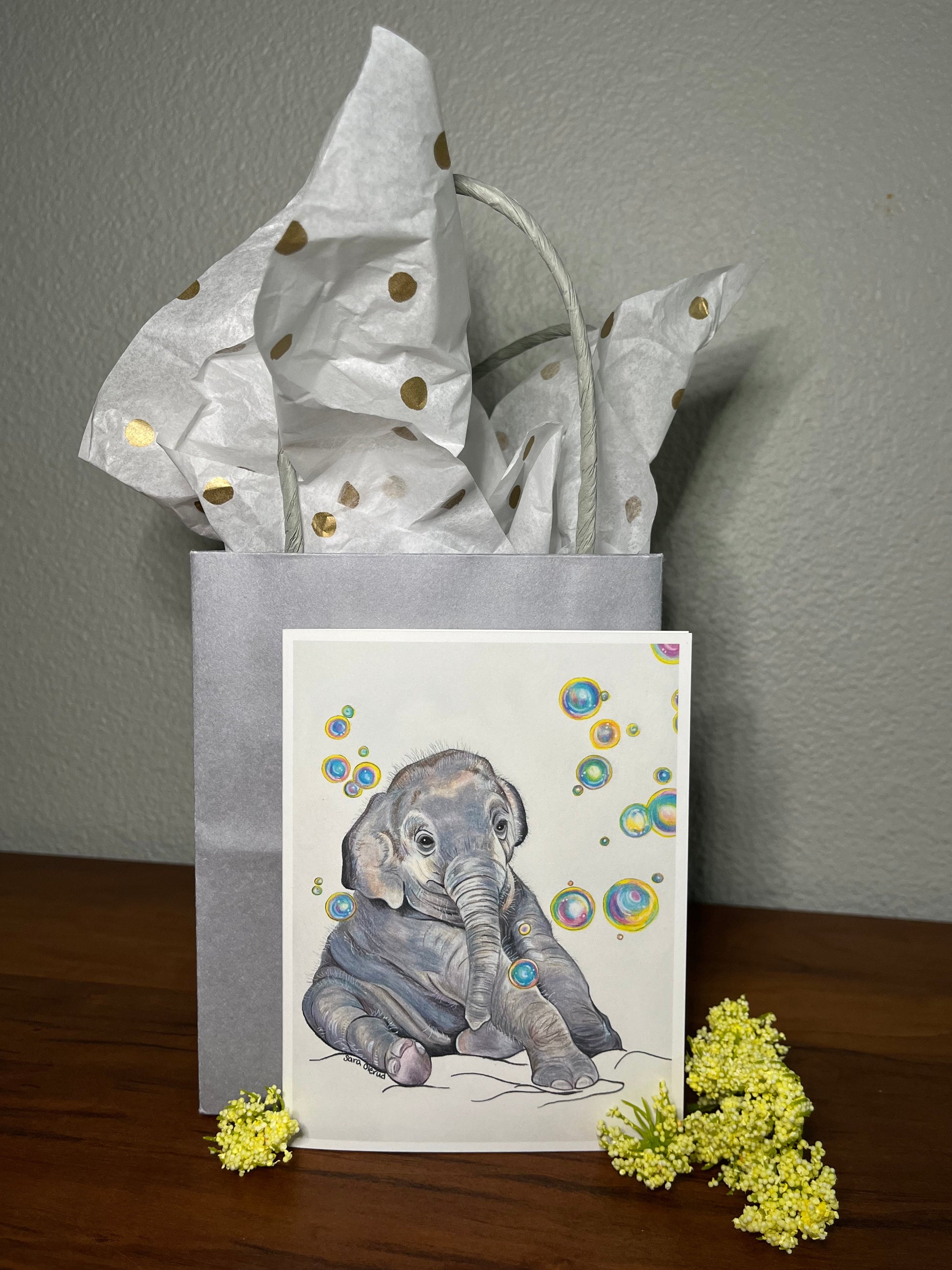 Gray gift bag with polka dot paper, card with elephant illustration, and yellow flowers on a wooden surface.