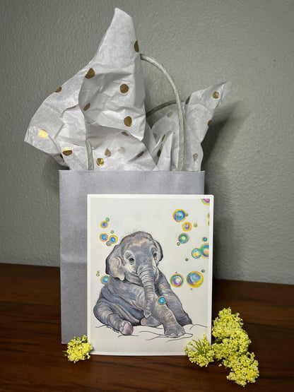 Gray gift bag with polka dot paper, card with elephant illustration, and yellow flowers on a wooden surface.