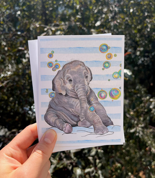 A hand holds a small art print or card featuring a watercolor-style baby elephant sitting on a blue-and-white striped background with colorful bubble accents, photographed outdoors in front of blurred green foliage.