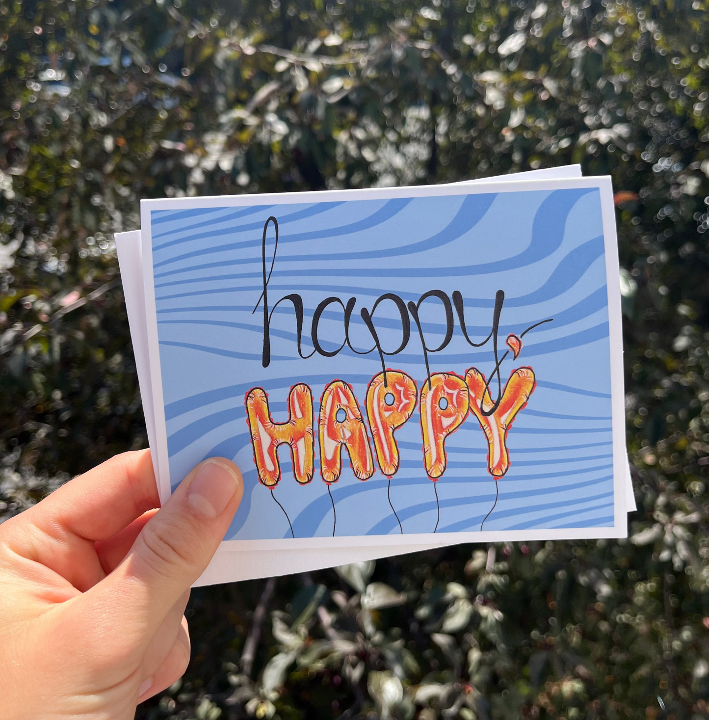 A hand holds a blue greeting card outdoors with blurred green foliage in the background. The card has wavy blue stripes and the words 'happy HAPPY' in black and orange lettering.