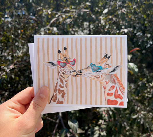 A hand holds a square art print featuring two giraffes wearing colorful sunglasses against a background of blurred green foliage. The print has vertical beige stripes on a white border.