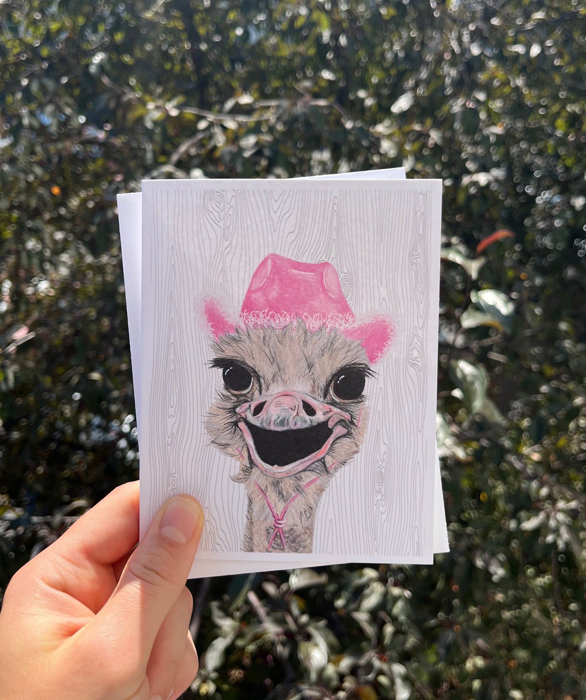 A hand holds a white art print featuring a stylized ostrich illustration with a pink hat, against a blurred outdoor background of green foliage.