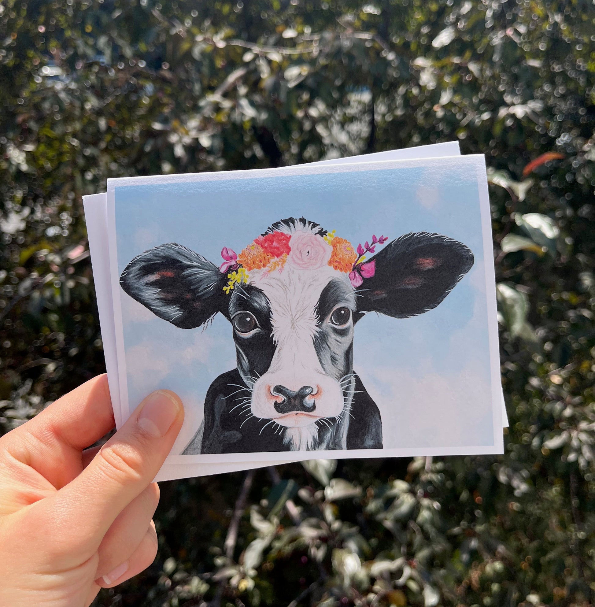 A hand holds a small printed card or postcard featuring a black-and-white cow with a colorful flower crown against a light blue background, with blurred green foliage behind it.