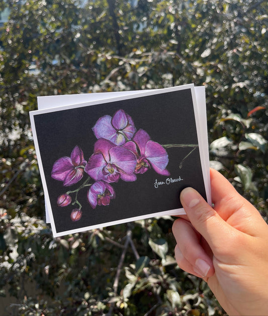 A hand holds a stack of art cards outdoors, with the top card showing a detailed drawing of purple orchids on a black background. The background is a blurred natural green foliage.