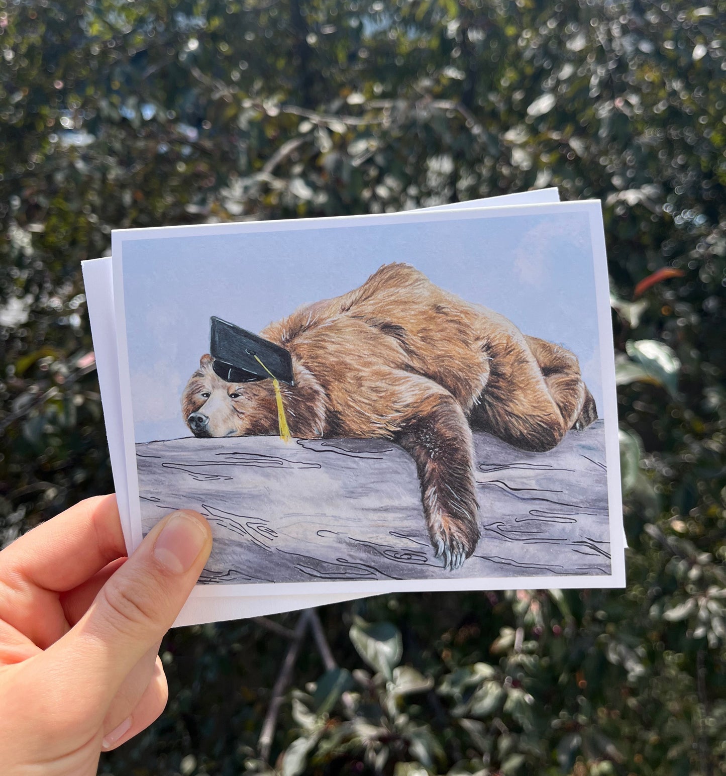 A hand holds a printed card featuring a sloth wearing a graduation cap, with a blurred outdoor background of green foliage.