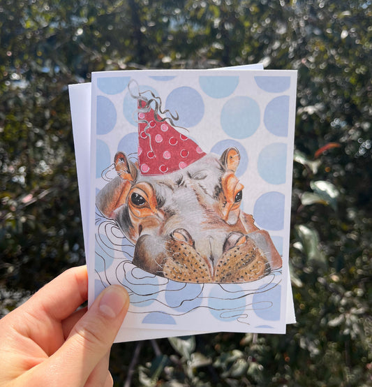 A hand holds a square art print or greeting card outdoors, showing an illustration of two hedgehogs wearing red party hats against a background of blue polka dots and water. The background of the photo is blurred green foliage.