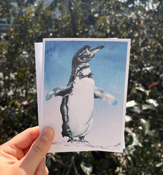 A hand holds a printed card featuring a penguin illustration against a background of blurred outdoor foliage.