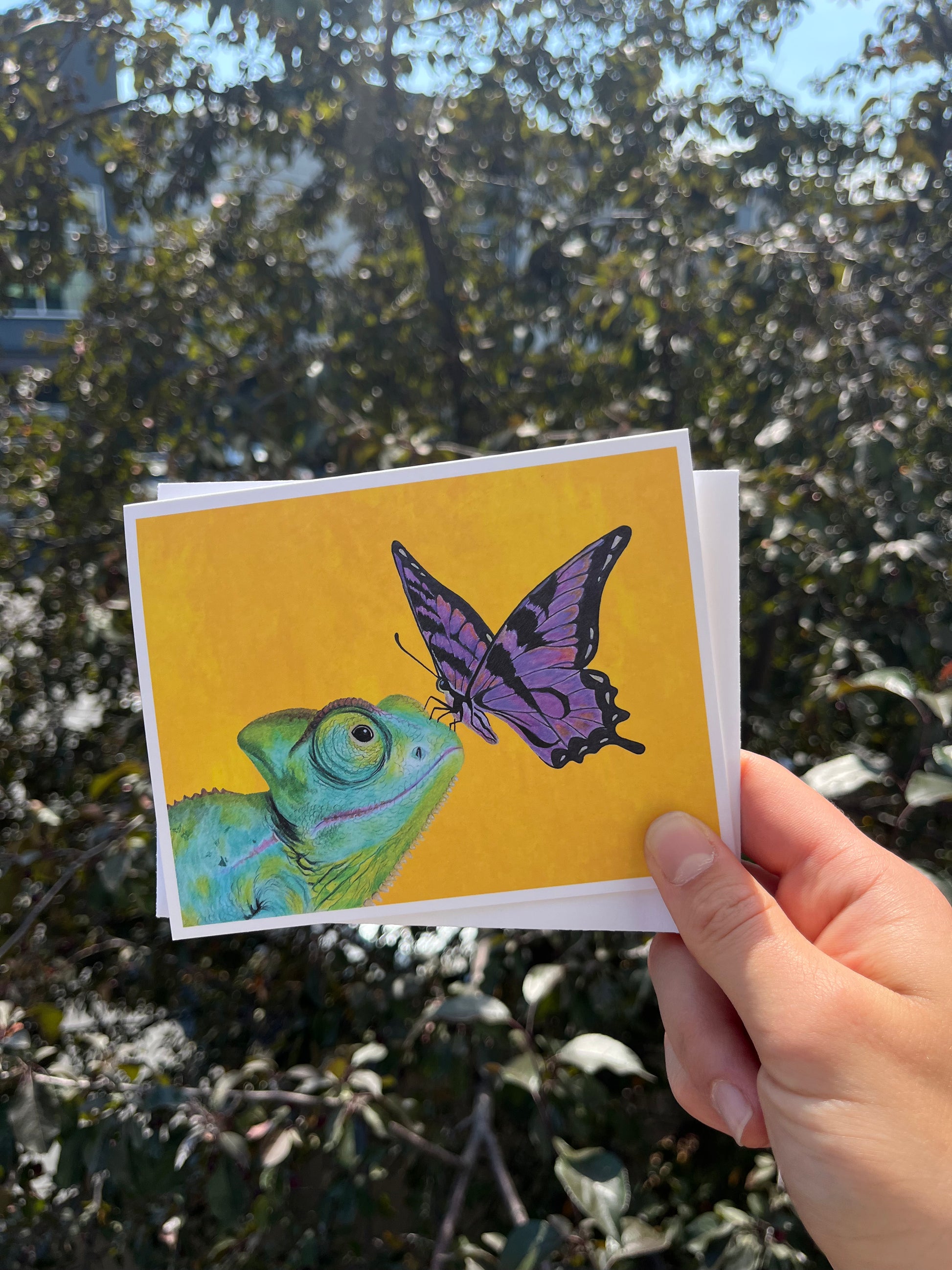 A hand holds a square art print featuring a green chameleon and a purple butterfly on a bright yellow background, with a real outdoor garden and trees blurred in the background.