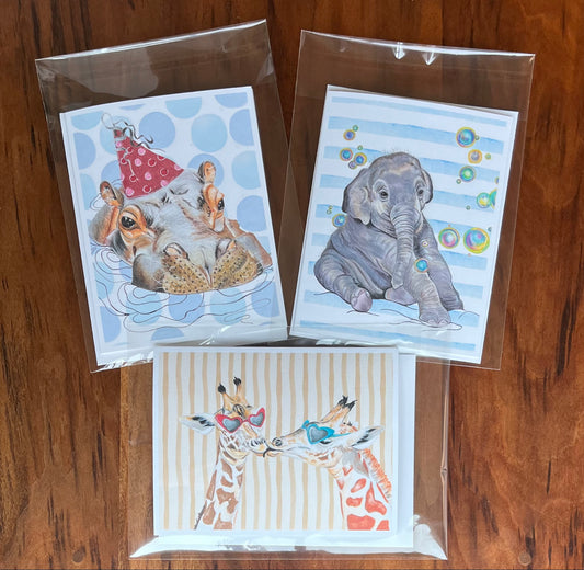 Three small art cards in clear plastic sleeves, arranged on a wooden surface. The top-left card shows rabbits in a blue polka-dot setting with a red hat.
