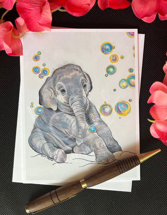 Card with a drawing of a baby elephant surrounded by colorful bubbles, placed on a black surface with pink flowers.